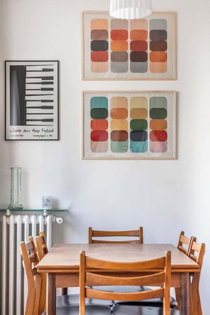 A dining area with a wooden table and chairs, a radiator, and two framed artworks on the wall.