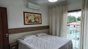 Ein Schlafzimmer mit Bett, Nachttisch, Klimaanlage, Fenster mit Vorhängen und einem Wandbild.