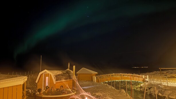 A wooden cabin with a hot tub and a covered area, surrounded by snow and a wooden fence.