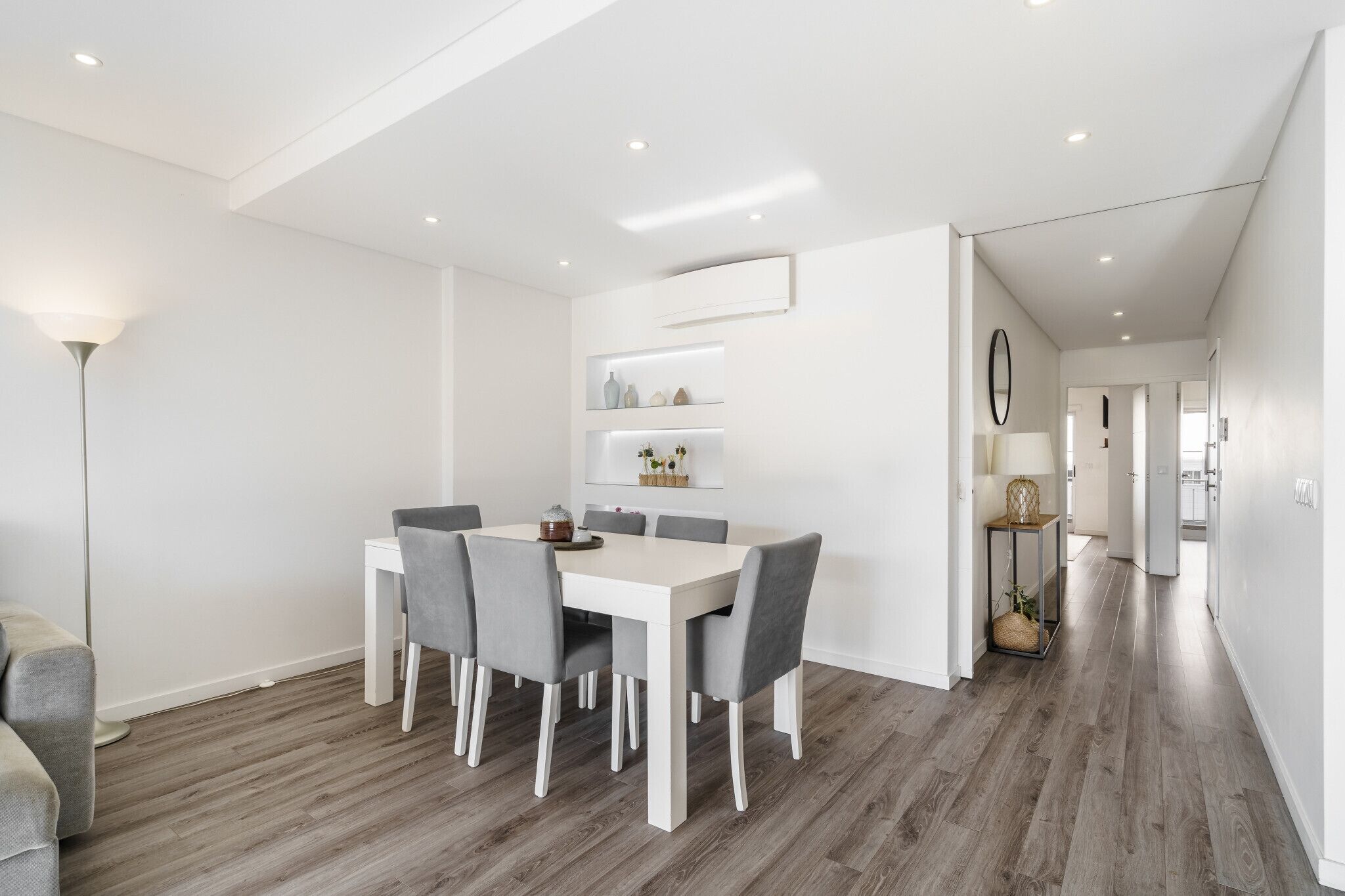 Un espace repas moderne avec une table blanche et des chaises grises, un lampadaire et un couloir menant à d’autres pièces.
