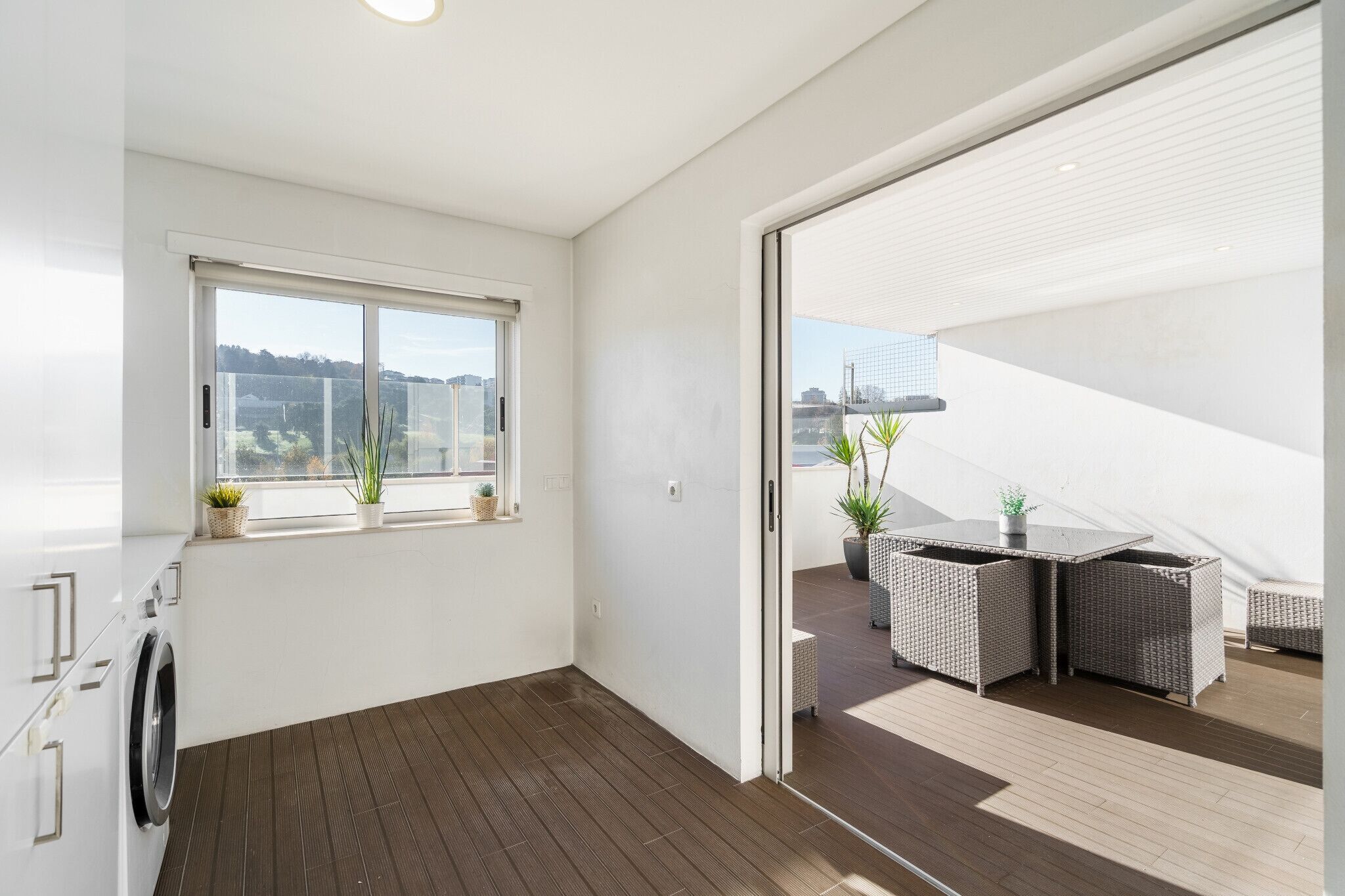 A modern apartment with a large window, a balcony with wicker furniture, and a washing machine.