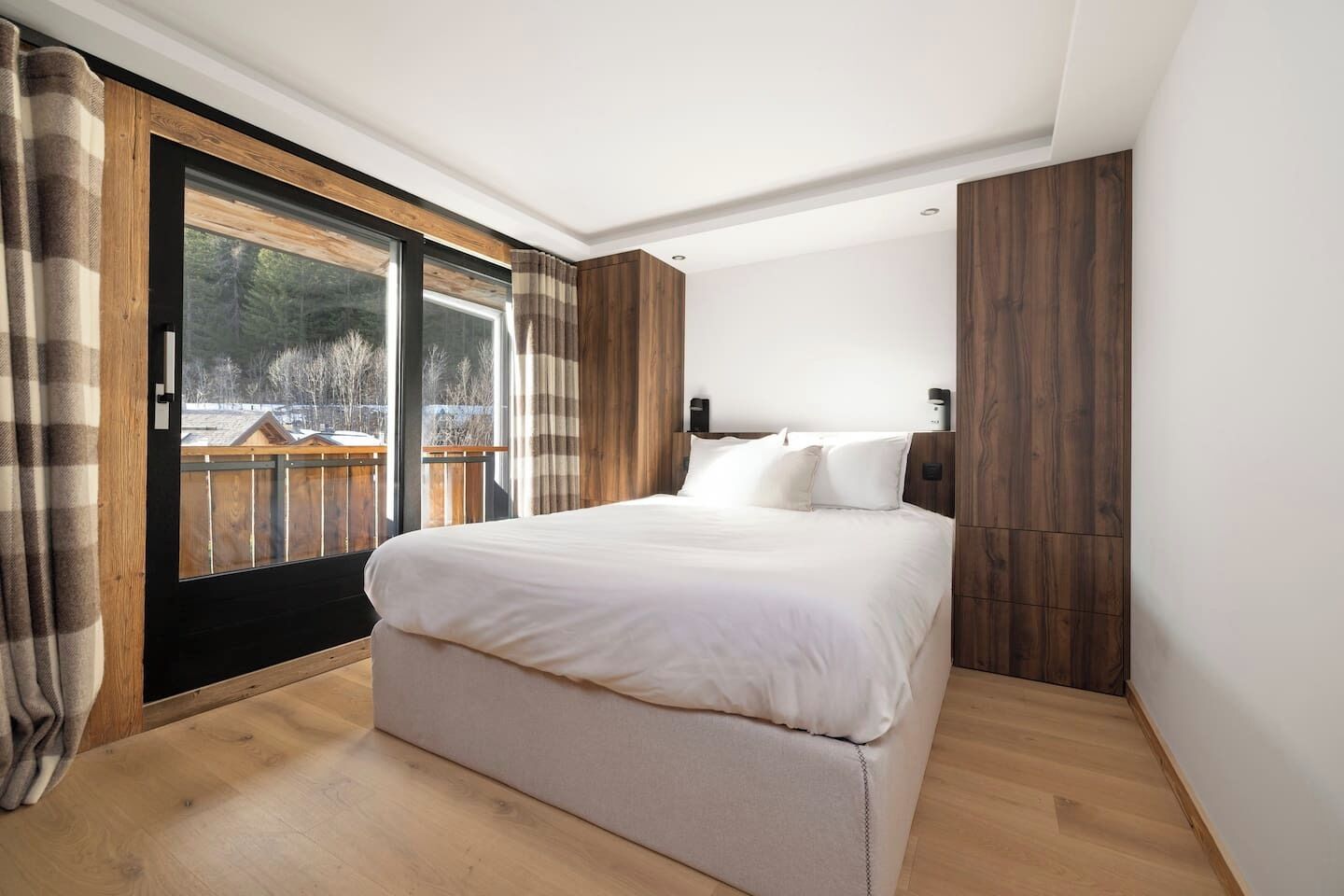 A bedroom with a large bed, wooden flooring, a sliding glass door leading to an outdoor area, and a view of trees and a wooden deck.