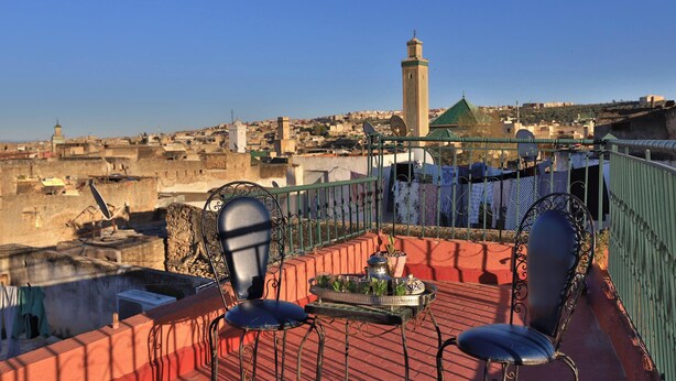 A rooftop terrace with two chairs, a tray with a plant, and a view of a cityscape including a prominent tower. - Spacious 5-bedroom riad in lovely Fès with AC and fitness room (Fès)