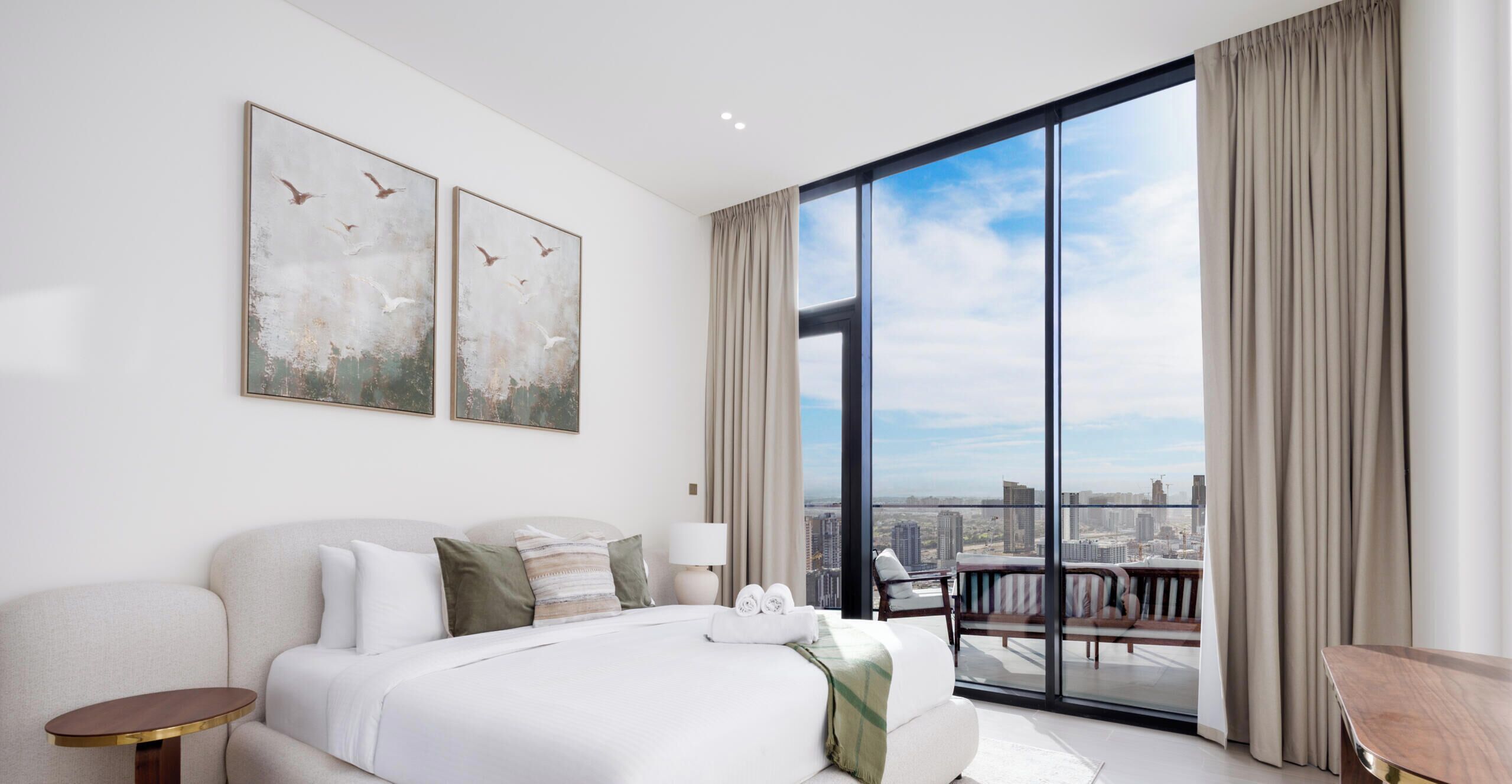 A modern hotel room with a large bed, a balcony with a city view, and two abstract paintings on the wall.