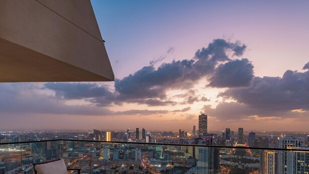 A cityscape view from a high-rise building with a glass balcony, featuring a sunset sky and illuminated buildings. - LUX  The Phantom Sky Pool Suite (Dubai)