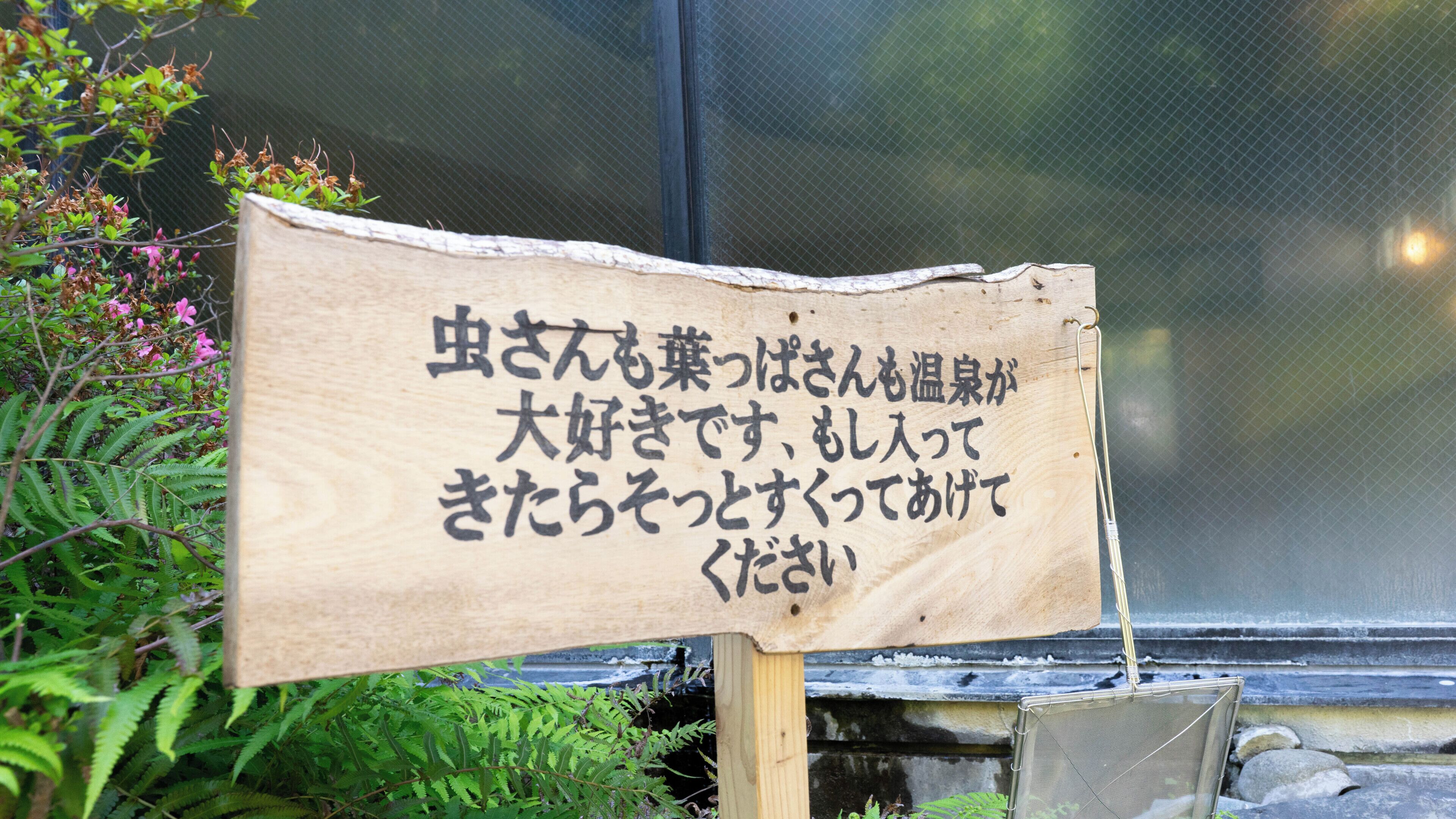 Un letrero de madera con texto japonés, rodeado de follaje y una cerca de malla.