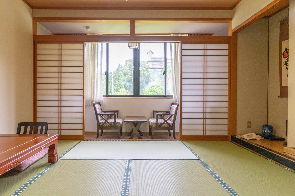 Une pièce de style japonais traditionnel, avec un sol recouvert de tatamis, une table basse, deux chaises et une porte coulissante donnant sur une fenêtre offrant une vue sur la verdure.