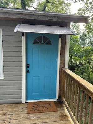 A blue door with a small window above it, a doormat with a message, and a wooden deck with a railing.