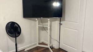 A room with a flat-screen TV mounted on a white stand, a standing fan, a door, and a trash can. - Private Studio 12min from. Mercedes Benz/Downtown (Atlanta)