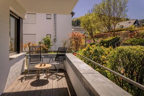 Un balcon avec une table et des chaises, une vue sur un jardin avec différentes plantes et arbres, et un bâtiment en arrière-plan.