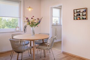 Un coin repas comprenant une table ronde, quatre chaises, un vase avec des fleurs, un luminaire suspendu et une étagère avec des tasses.