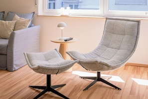 A modern living room with a grey sofa, two grey armchairs, a small round wooden side table with a lamp, and a window with a view of buildings. - 5 minutes from the Green Hill Bikepark and the Hunau slope Family apartment! (Schmallenberg)