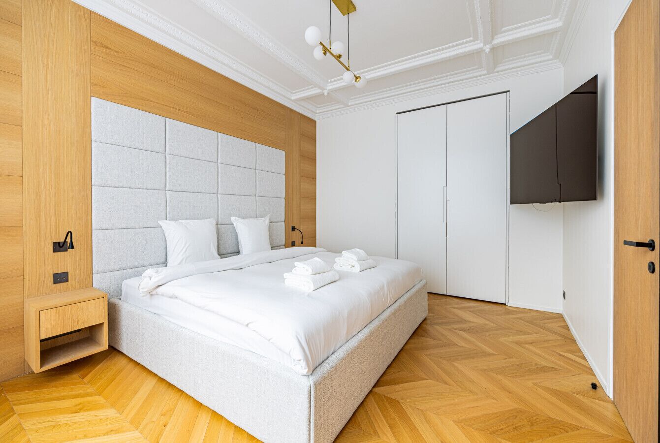 Ein modernes Schlafzimmer mit einem großen Bett, einem Holz-Kopfteil, einer integrierten Nachttischschublade, einem flachbildfernseher an der Wand und einem Holzboden mit Fischgrätmuster.