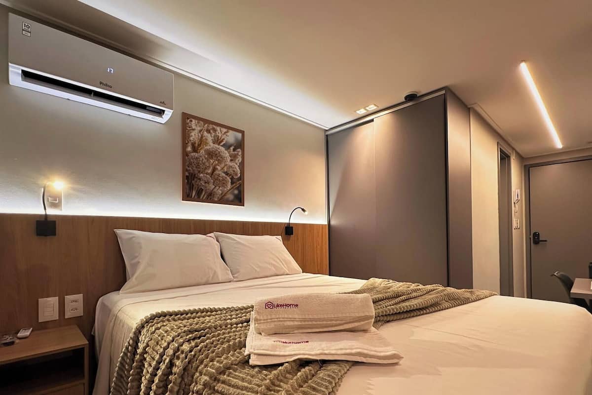 Une chambre d’hôtel comprenant un lit, une table de chevet, un climatiseur fixé au mur, une lampe fixée au mur, une œuvre d’art encadrée et une porte.