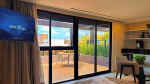 A modern room with a large sliding glass door leading to an outdoor patio, a flat-screen TV mounted on the wall, a dining area with a table and chairs, and a kitchenette with a coffee machine. - HIT Cabo Branco II- LikeHome Hospedagens (João Pessoa)
