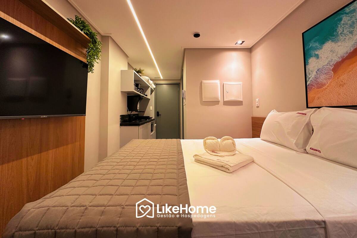 Une chambre d’hôtel comprenant un lit, un téléviseur à écran plat, une étagère murale avec une plante, un tableau représentant une plage et un panneau indiquant « LikeHome ».