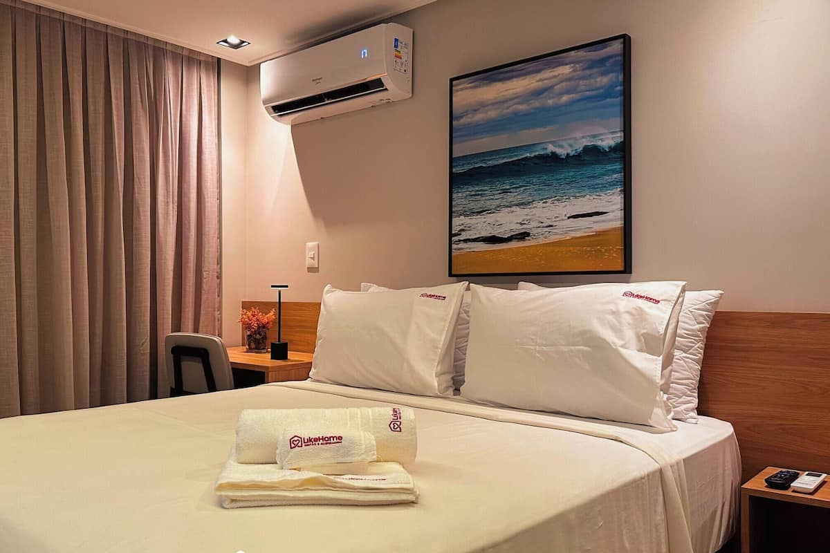 Une chambre d’hôtel avec un lit, un bureau, un climatiseur et un tableau représentant une vue sur la plage.