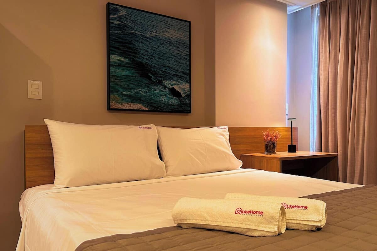 Une chambre d’hôtel avec un lit, un tableau représentant l’océan, une table de chevet avec un vase et une fenêtre avec des rideaux.