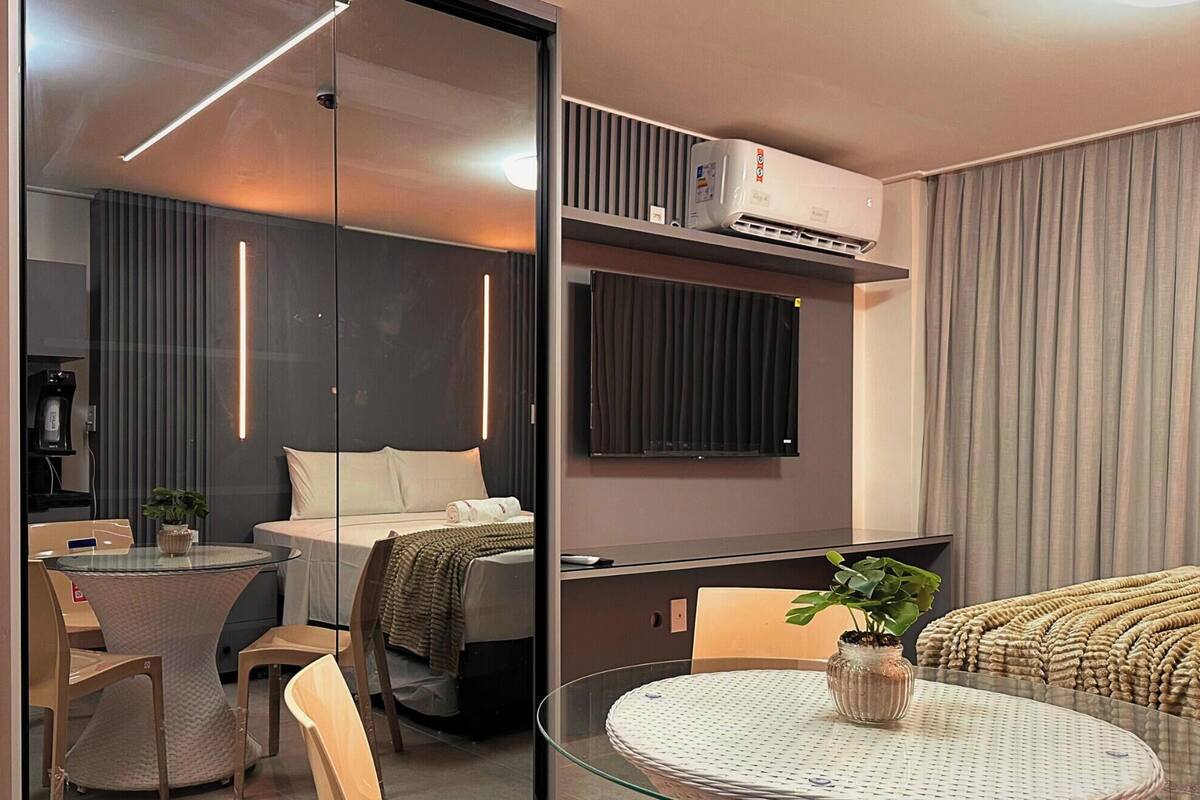 Une chambre d’hôtel moderne comprenant un lit, une table en verre, une chaise, une petite table avec un vase et une plante en pot.