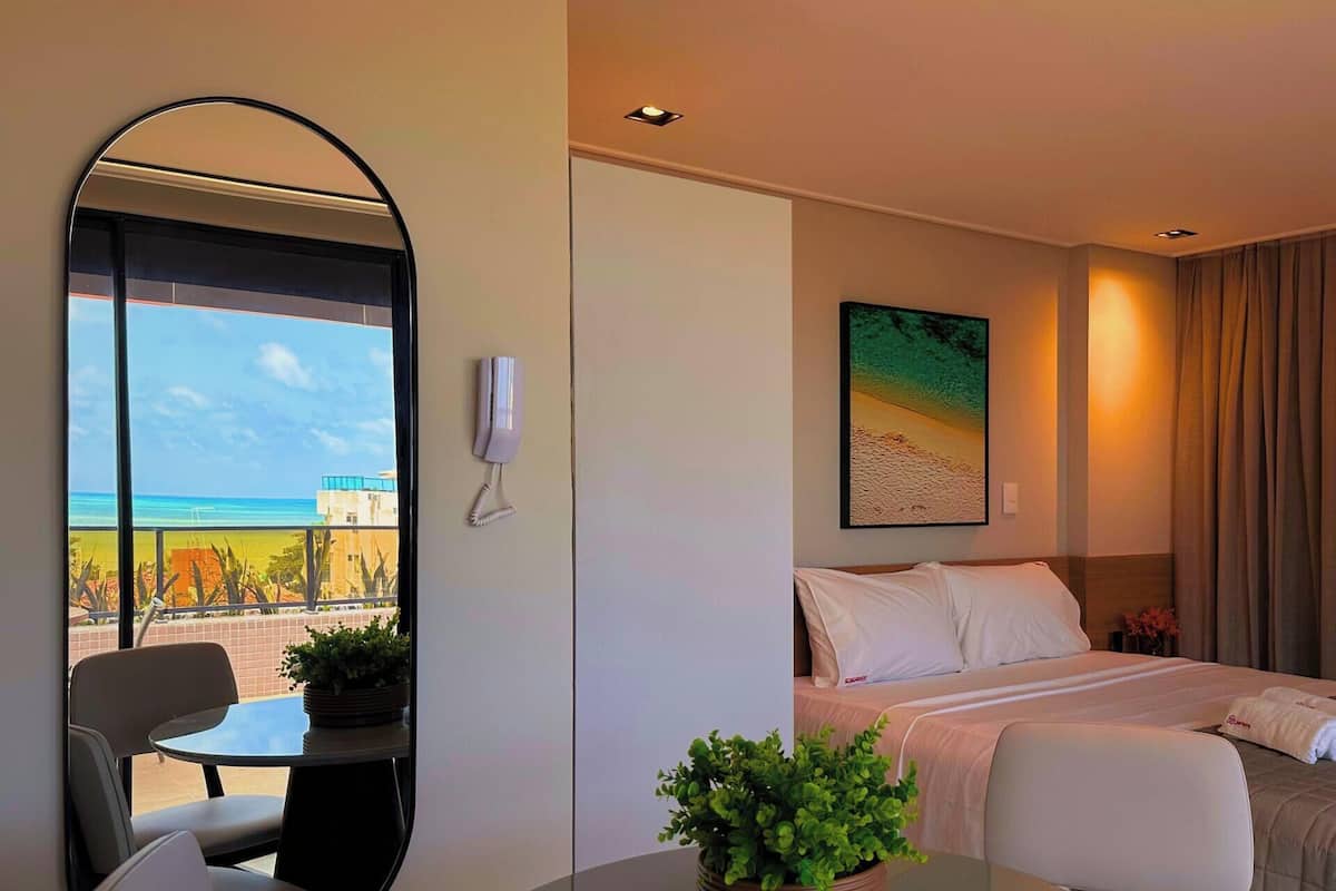 Une chambre d’hôtel avec un lit, un bureau, une chaise, une plante en pot, un tableau et une vue sur la plage.