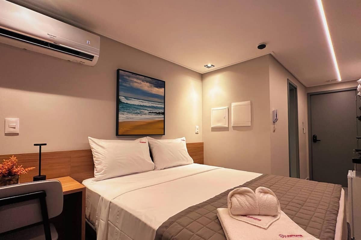 Une chambre d’hôtel avec un lit, un bureau, un climatiseur, un tableau représentant une plage et une porte.