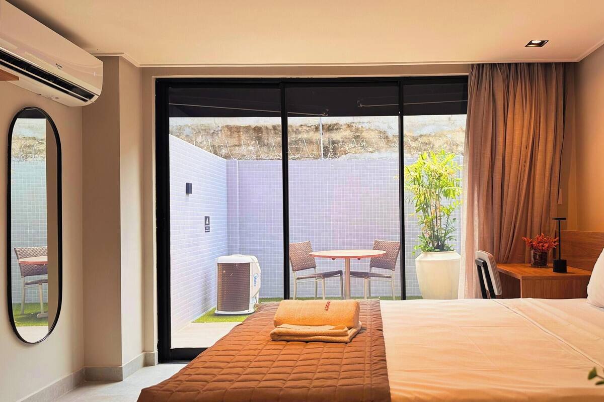 Une chambre d’hôtel avec un grand lit, une petite table avec des chaises, une plante en pot et une vue sur un patio extérieur avec une table et des chaises.
