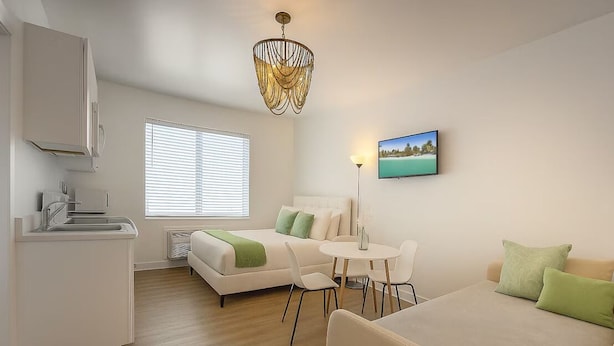 Quarto moderno e bem iluminado, com cozinha compacta, cama com almofadas verdes, área de refeições e uma janela grande com persianas.