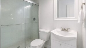 Casa de banho com chuveiro de cabine de vidro, sanita branca e lavatório branco com espelho em cima.