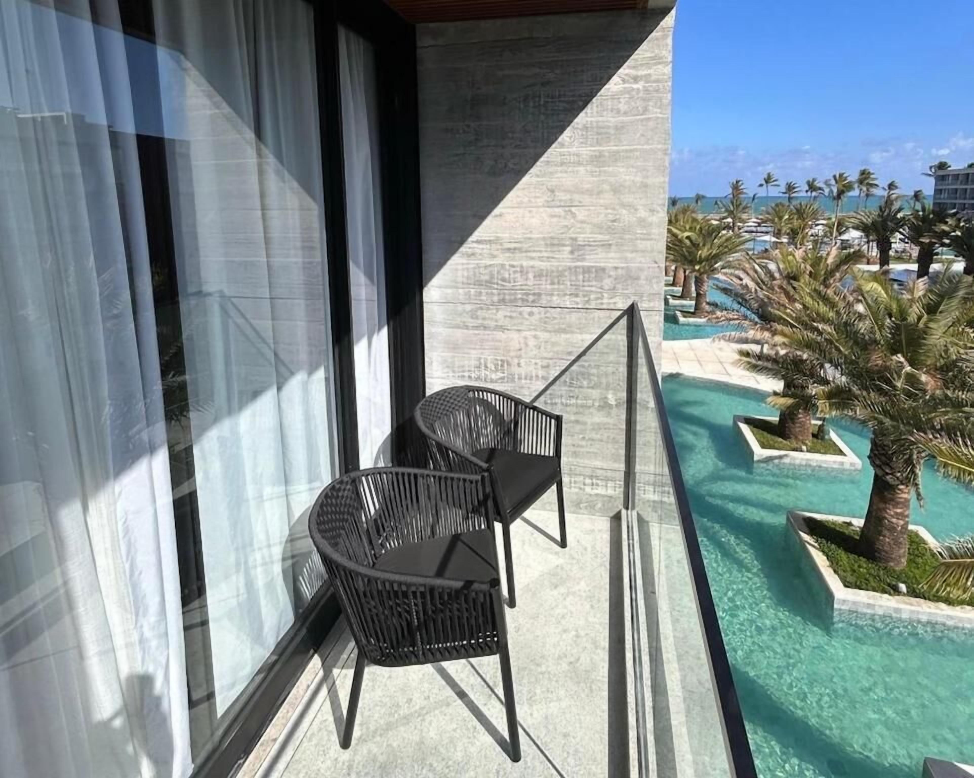 Un balcon avec deux chaises noires, un mur en béton et une vue sur un espace piscine agrémenté de palmiers et d’un bâtiment en arrière-plan.