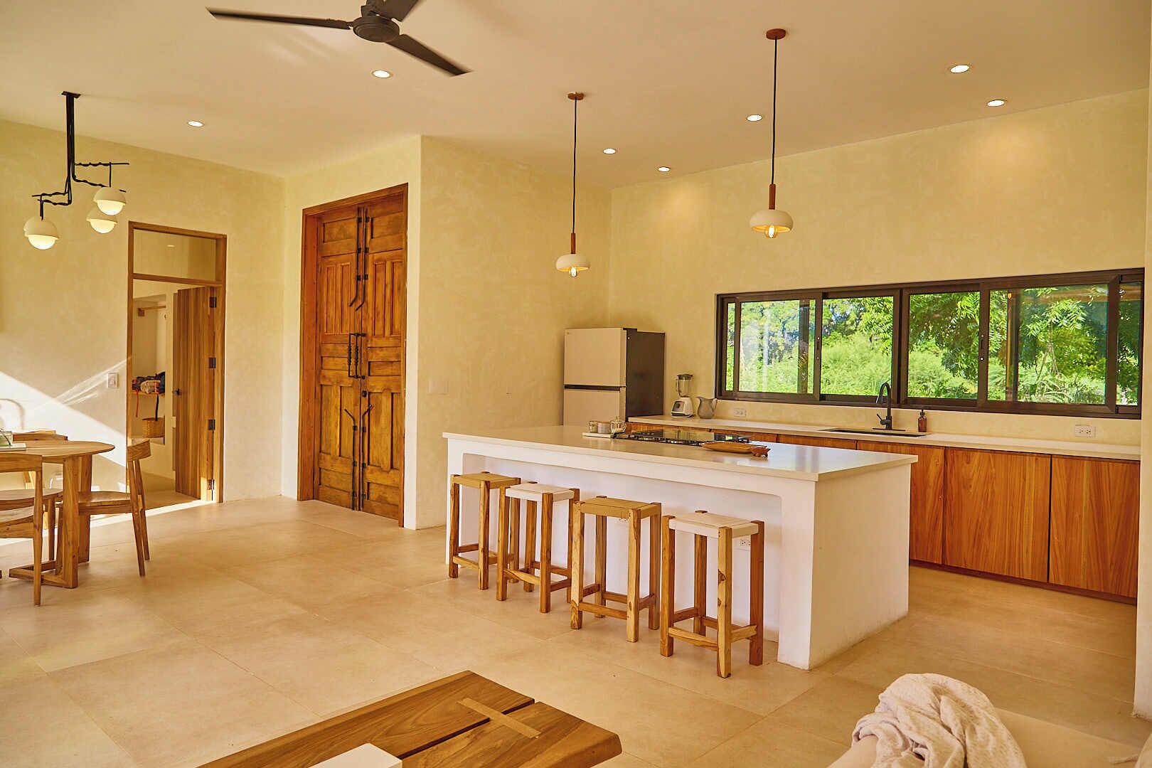 Una cocina moderna con una isla central, taburetes de madera, un comedor con mesa y sillas, y un ventanal con vistas a la vegetación.