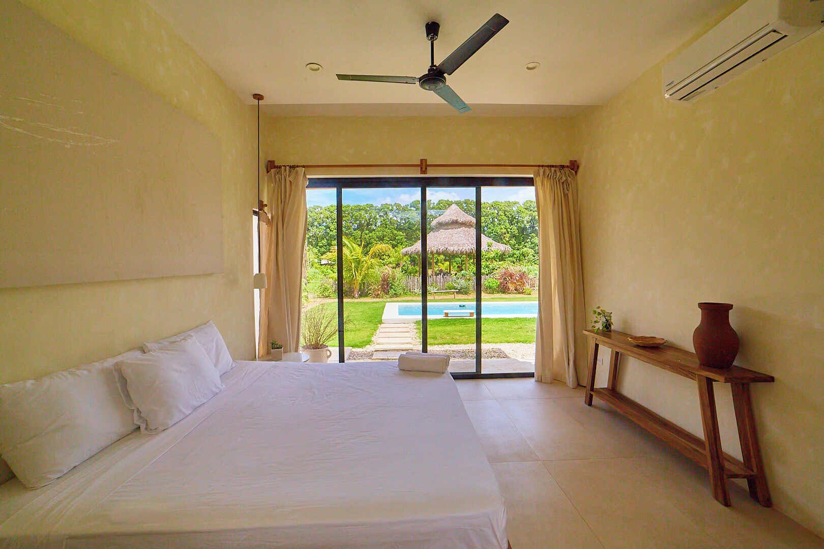Un dormitorio con cama, ventilador de techo, ventana con cortinas, vista a una piscina y jardín, y una mesita con un florero.