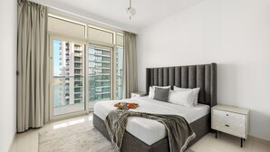 A modern bedroom with a large bed, a nightstand, and a view of high-rise buildings through floor-to-ceiling windows. - 2BR Beachfront Retreat with Palm Views (Dubai)