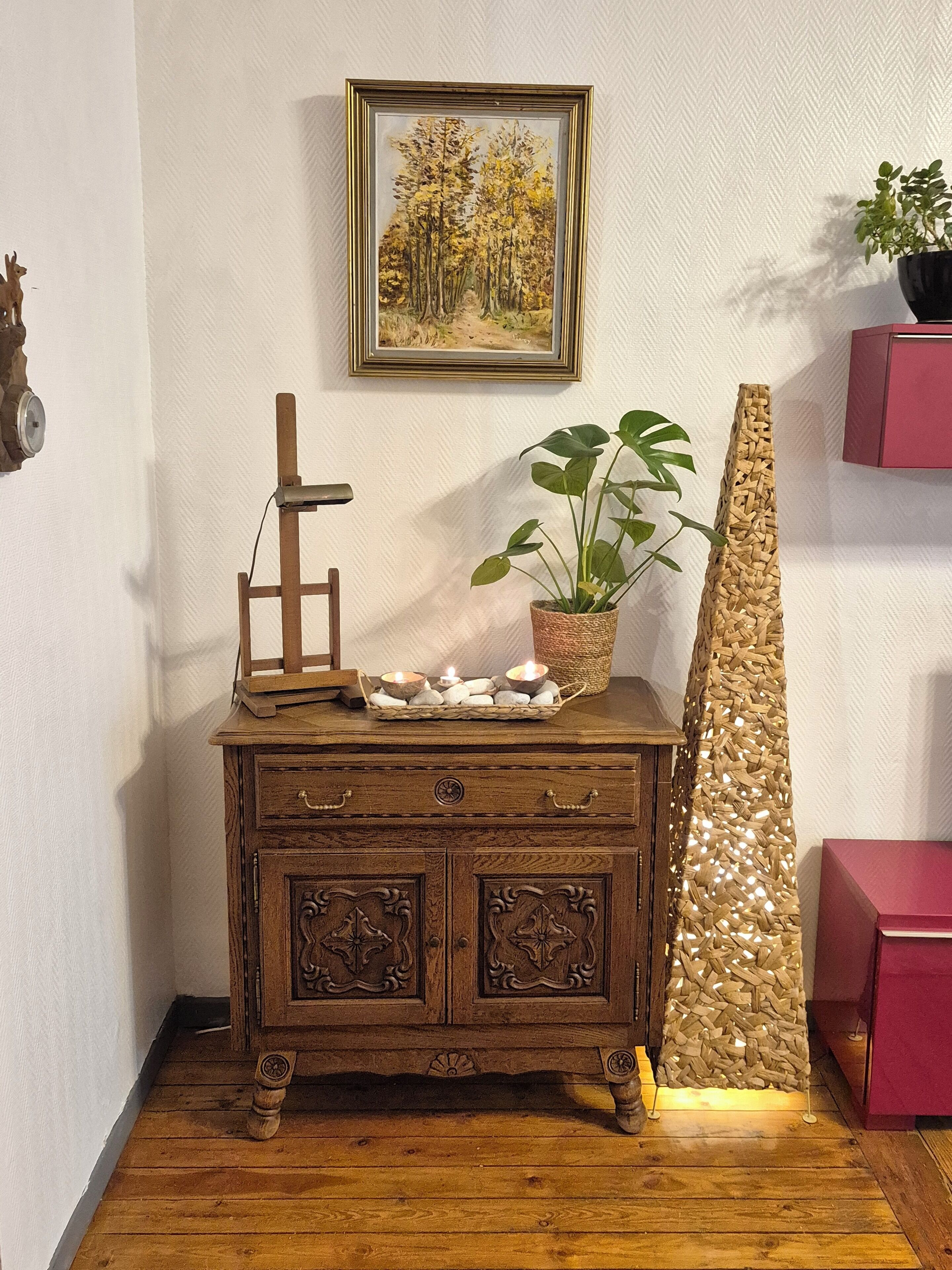 A wooden dresser with a framed painting, a potted plant, and a decorative wall piece.