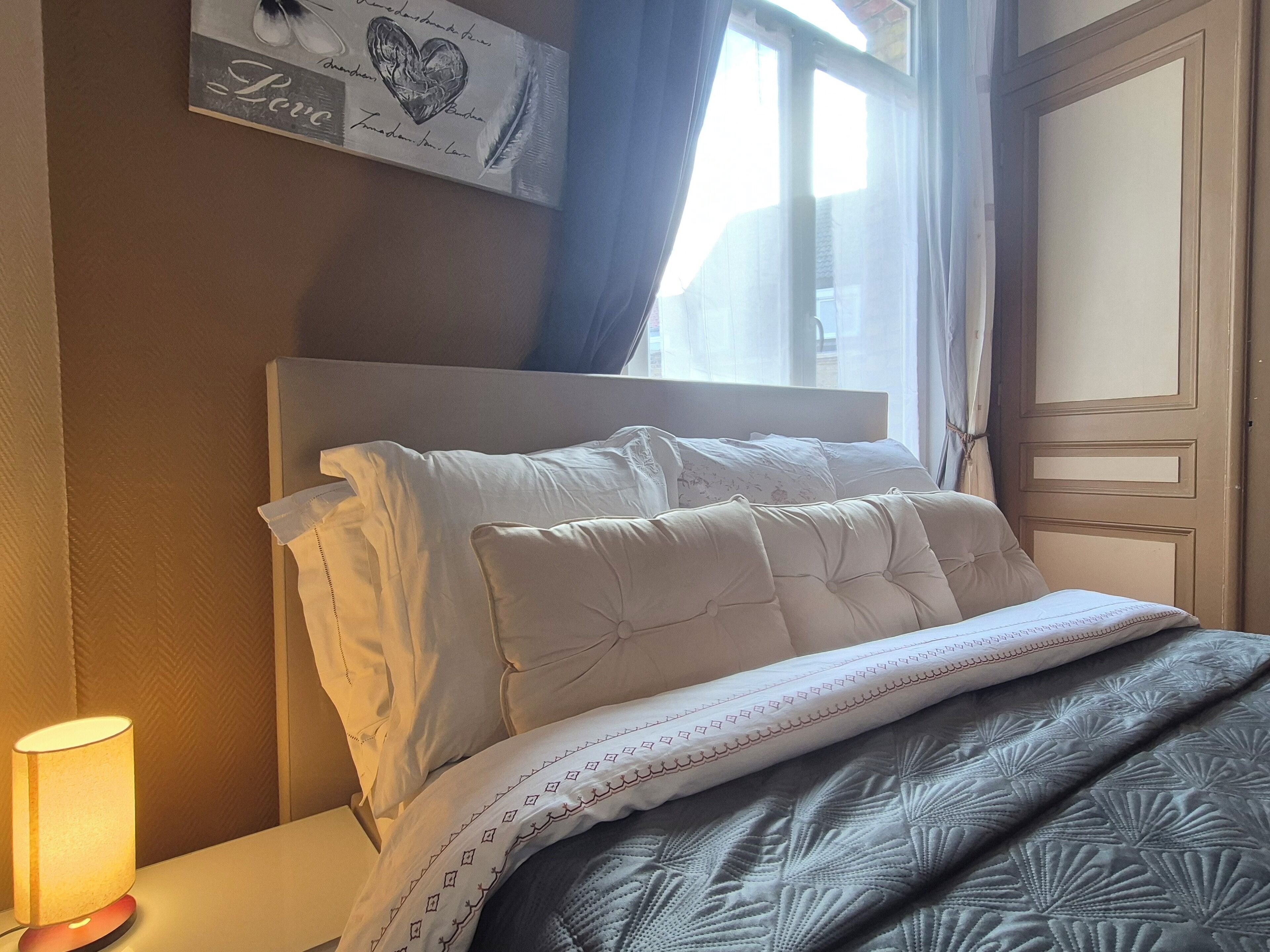 Una cama bien hecha con sábanas blancas y una colcha estampada, una lámpara de noche y una decoración en la pared con un corazón y la palabra 'Love'.