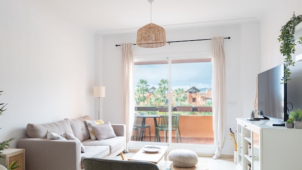 A modern living room with a sofa, a coffee table, a TV, and a balcony with a view of palm trees.