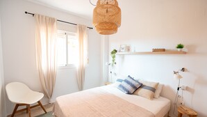 A bedroom with a bed, a chair, a bedside table, a lamp, a shelf with a plant, and a window with curtains.