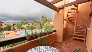 A balcony with a view of a pool and palm trees, a spiral staircase, and a table with chairs.
