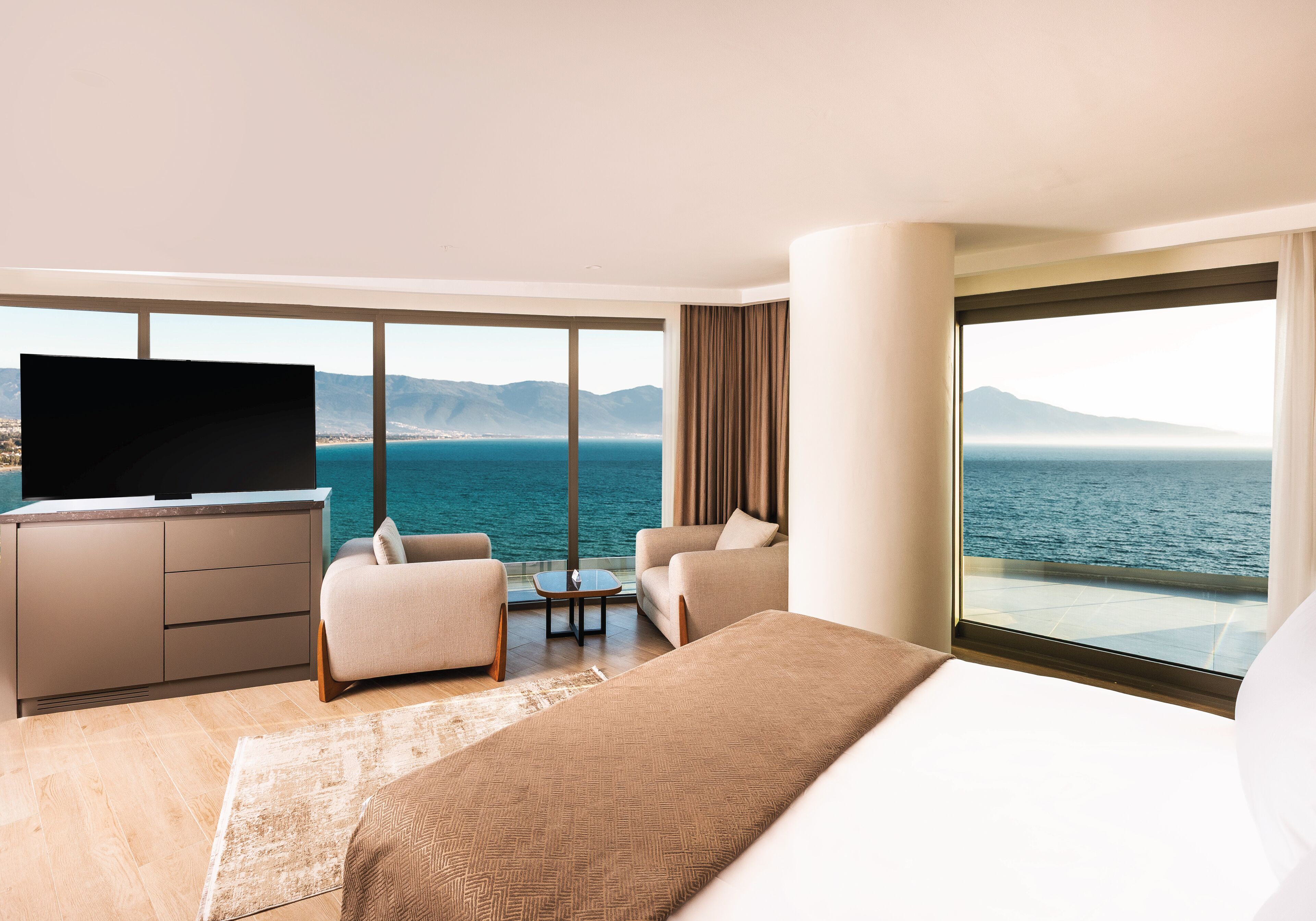 Habitación de hotel con una cama grande, televisión, dos sillones, una mesita y vistas al mar y a las montañas.