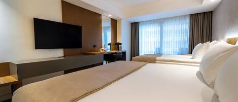 A hotel room with a large bed, a desk with a chair, a flat-screen TV mounted on the wall, and a window with curtains.