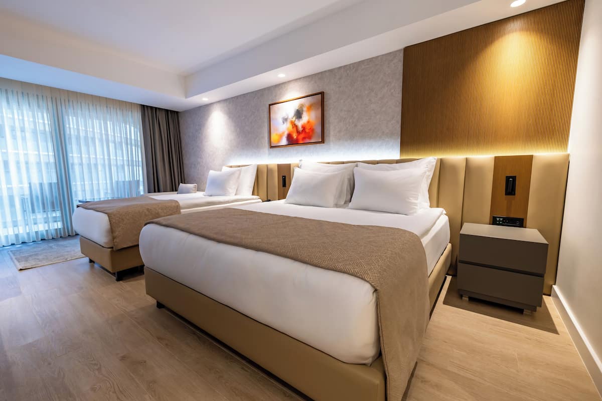 A modern hotel room with a large bed, two bedside tables, a wall-mounted artwork, and a large window with curtains.