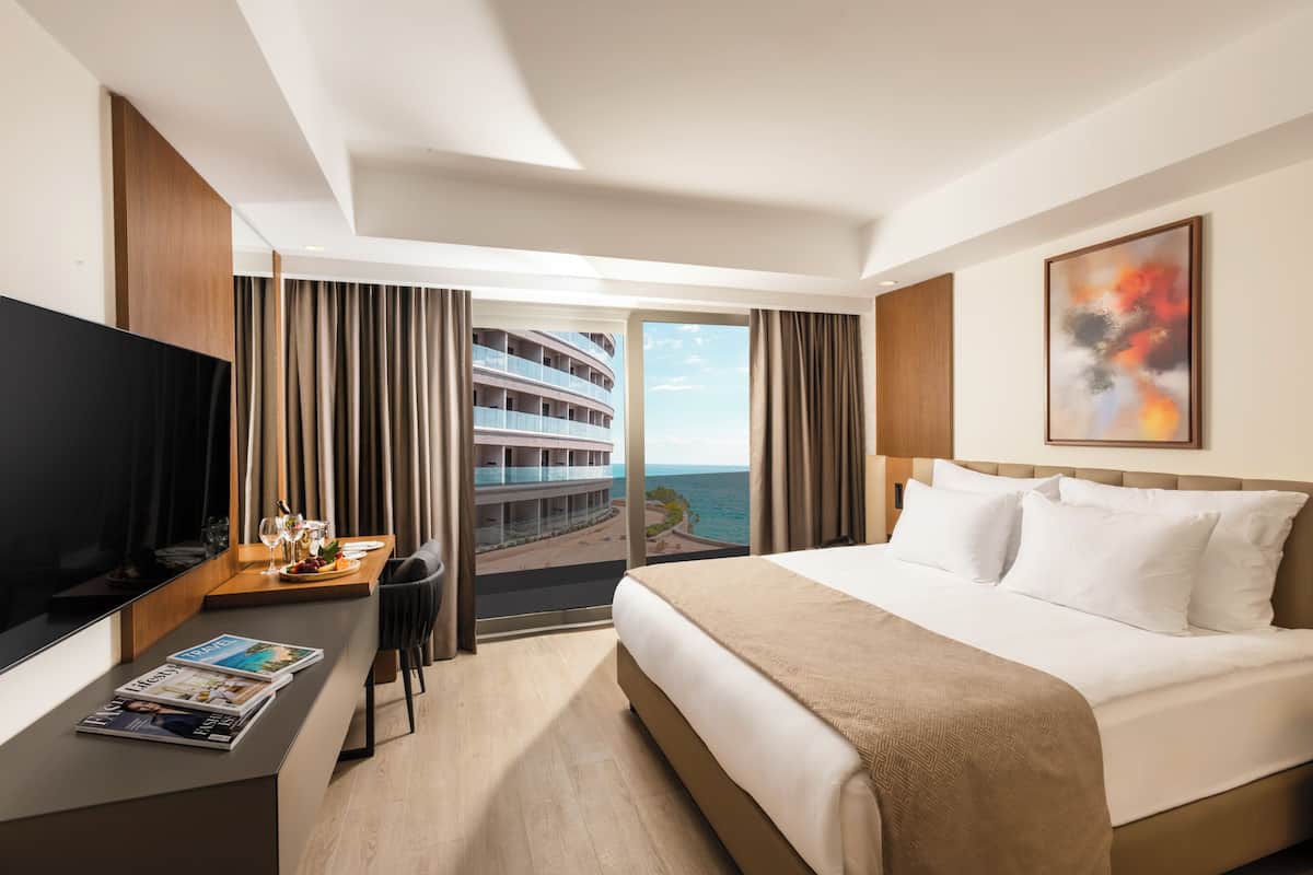 A hotel room with a bed, a TV, a desk with a magazine, and a view of a building and the sea.