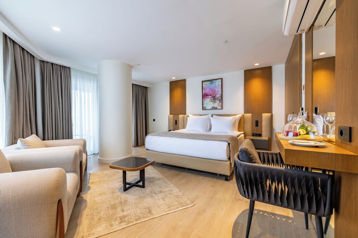 A modern hotel room with a bed, two armchairs, a small table, and a dining area with a chair.