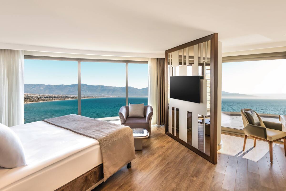 A hotel room with a bed, two chairs, a TV, and a large window with a view of the sea and mountains.