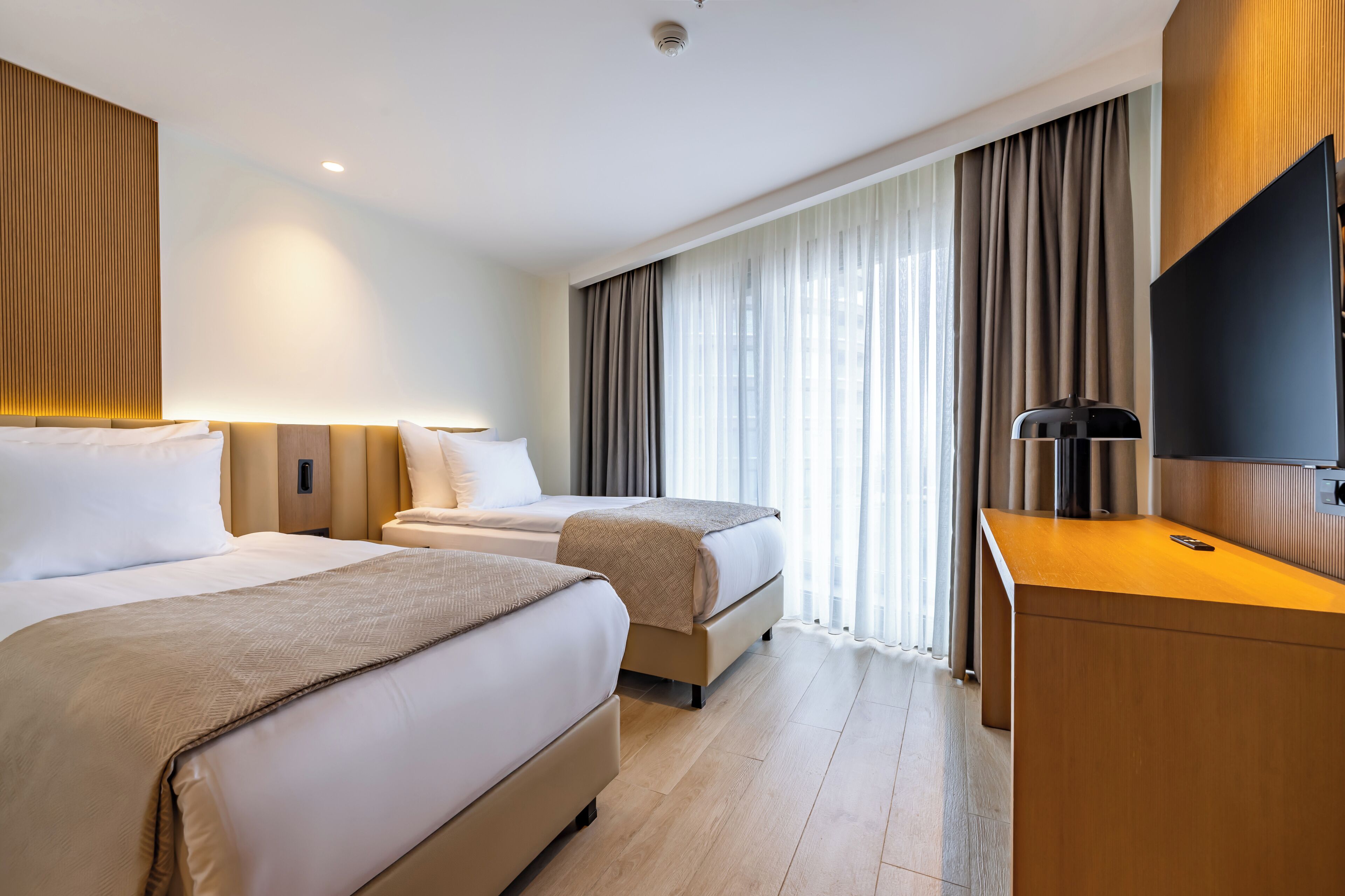 A hotel room with two beds, a desk, a flat-screen TV, and a large window with curtains.