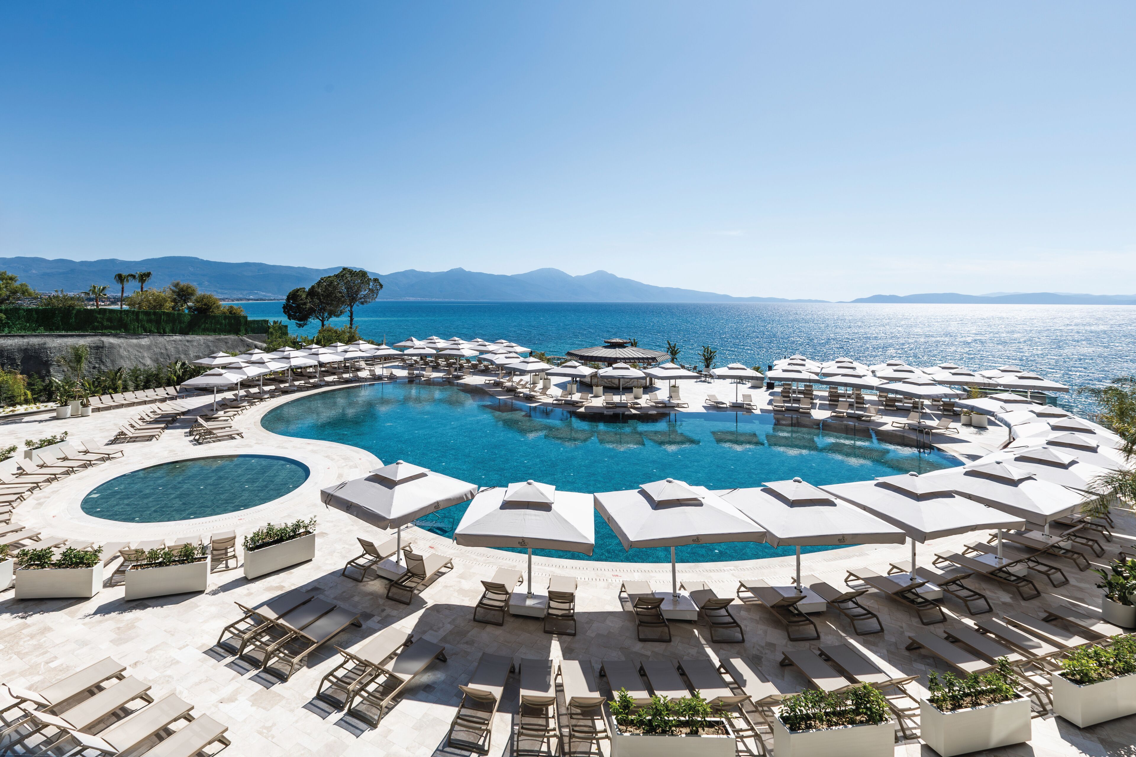 A circular infinity pool with lounge chairs and umbrellas, surrounded by a landscaped area with a view of the sea and mountains.