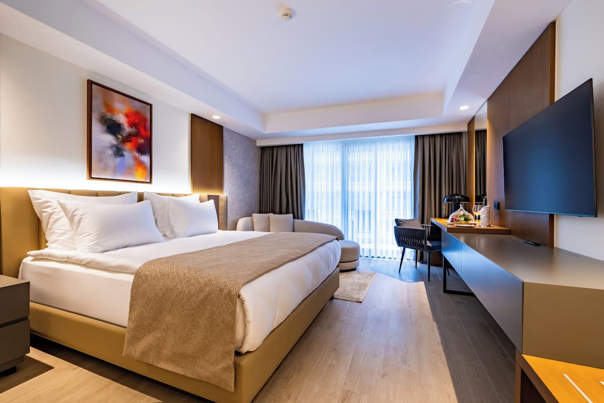 A modern hotel room with a large bed, a desk, a flat-screen TV, and a large window with curtains.