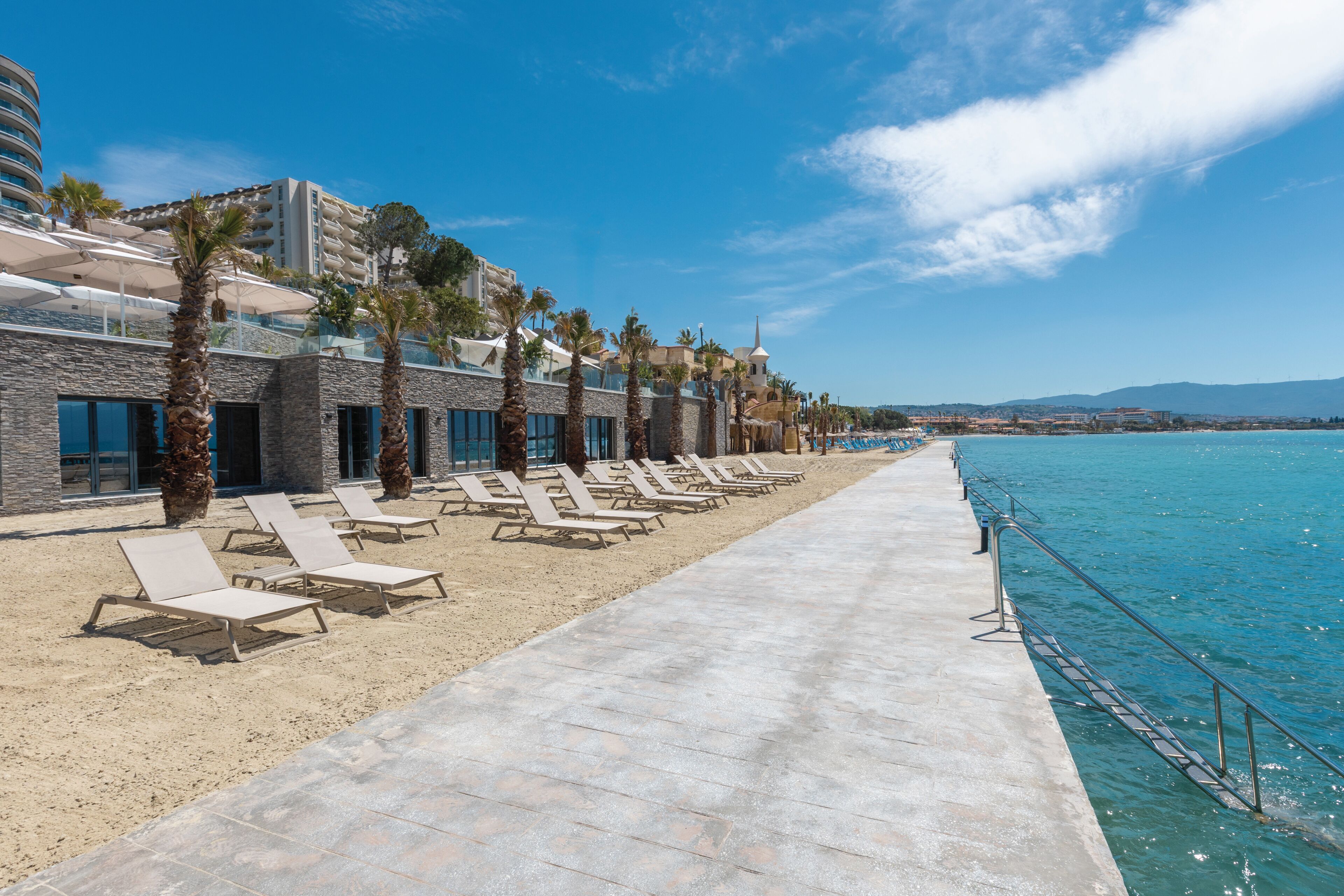 A beachfront hotel with sun loungers, palm trees, and a clear blue sky.