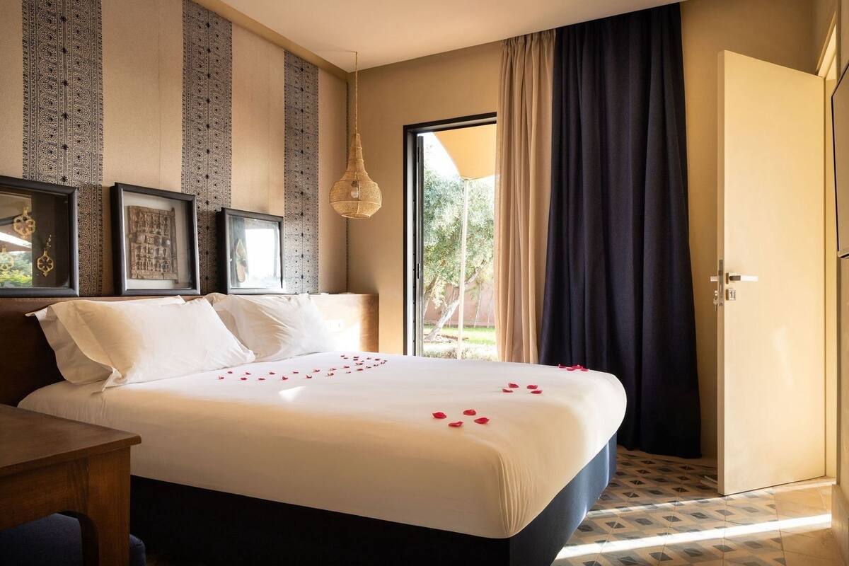 A hotel room with a bed, a wooden nightstand, a hanging light fixture, and a door leading to a balcony.