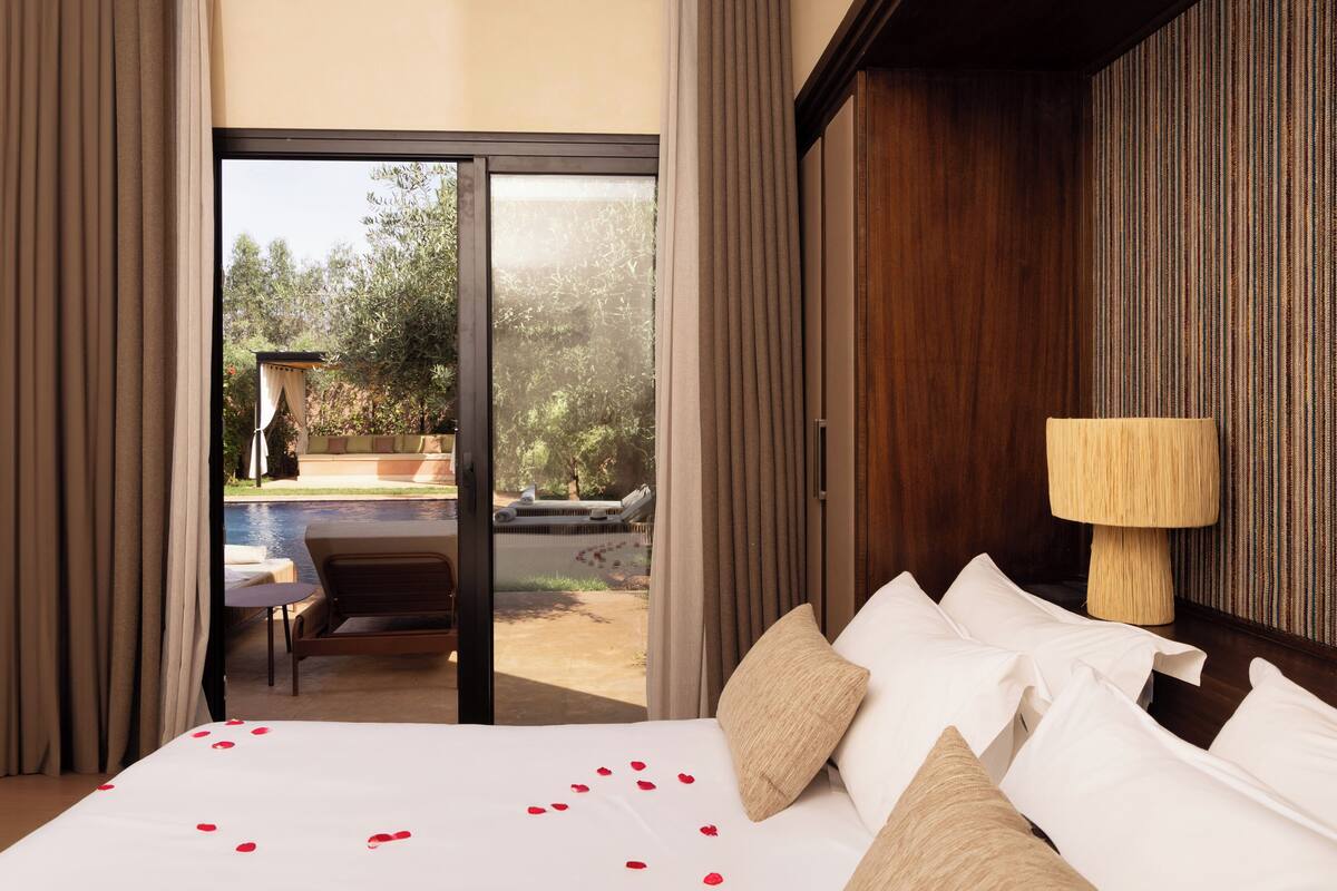 A hotel room with a bed, a lamp, and a view of a pool and garden.