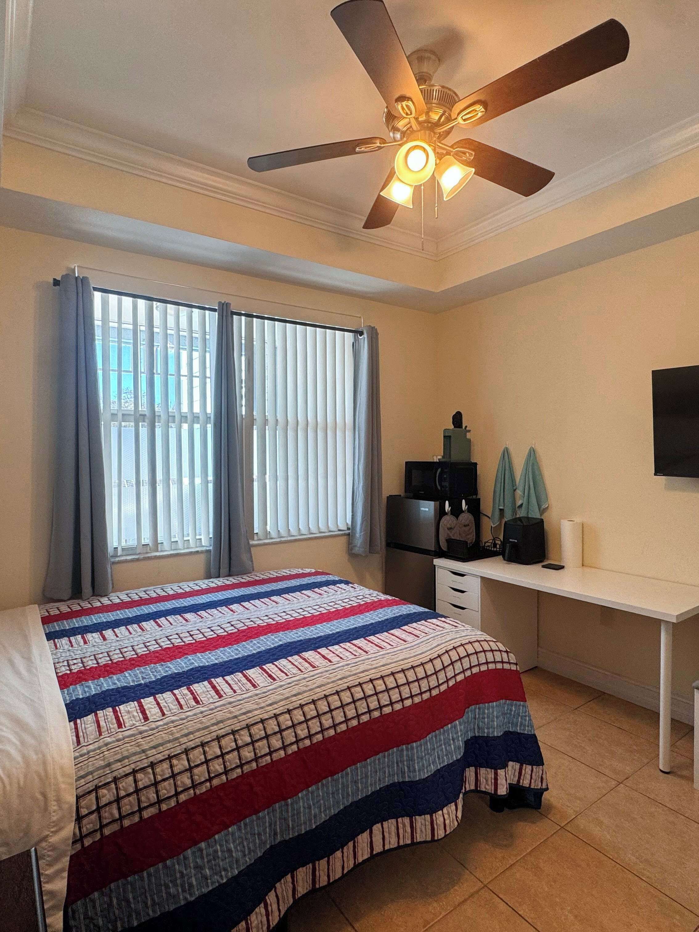 Quarto com cama, escrivaninha, ventoinha no teto, janela com cortinas e televisão na parede.